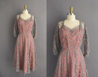 May include: A vintage lace dress with a blush pink bodice and a gray lace overlay. The dress has a fitted bodice and a full skirt. It features a sweetheart neckline and three-quarter sleeves.