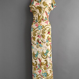 May include: A vintage ivory-colored dress with a floral pattern. The dress features a scoop neckline, short sleeves, and a fitted bodice that flows into a floor-length skirt with a slit. The floral design includes pink, blue, and green accents.