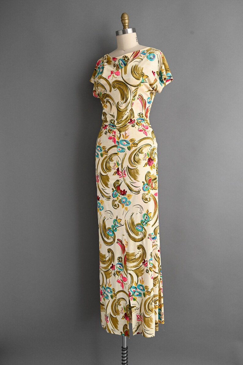May include: A vintage ivory-colored dress with a floral pattern. The dress features a scoop neckline, short sleeves, and a fitted bodice that flows into a floor-length skirt with a slit. The floral design includes pink, blue, and green accents.