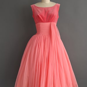 May include: A vintage coral-colored dress displayed on a mannequin. The sleeveless bodice is a darker shade of coral, with a cinched waist and a flowing, knee-length skirt. The dress has a classic, elegant design.