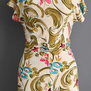 May include: Cream-colored dress with a floral pattern. The dress features short sleeves and a fitted waist. The design includes pink, blue, and yellow flowers, along with olive green swirls. The dress is displayed on a mannequin.