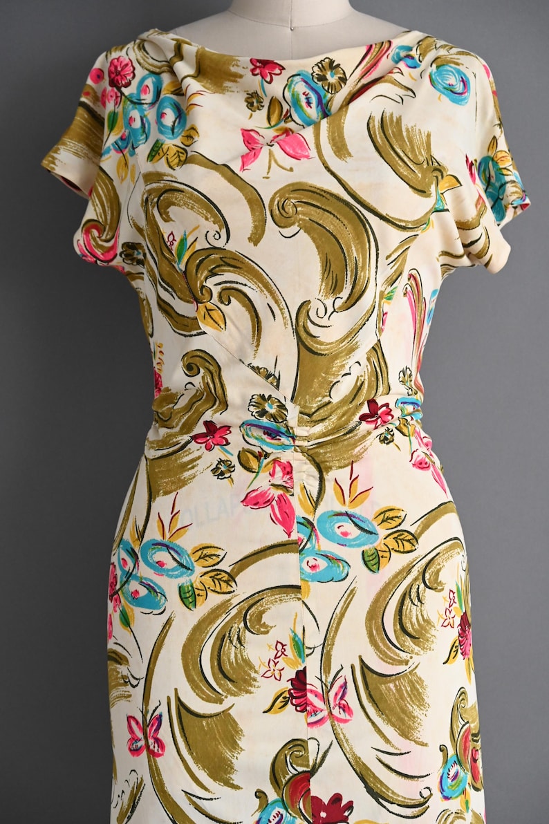 May include: Cream-colored dress with a floral pattern. The dress features short sleeves and a fitted waist. The design includes pink, blue, and yellow flowers, along with olive green swirls. The dress is displayed on a mannequin.