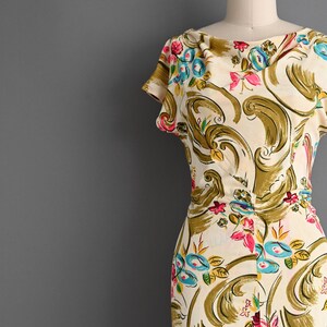 May include: A cream-colored dress featuring a cowl neckline and short sleeves, decorated with a repeating pattern of large olive green swirls, bright pink flowers, and small blue floral accents throughout the fabric.