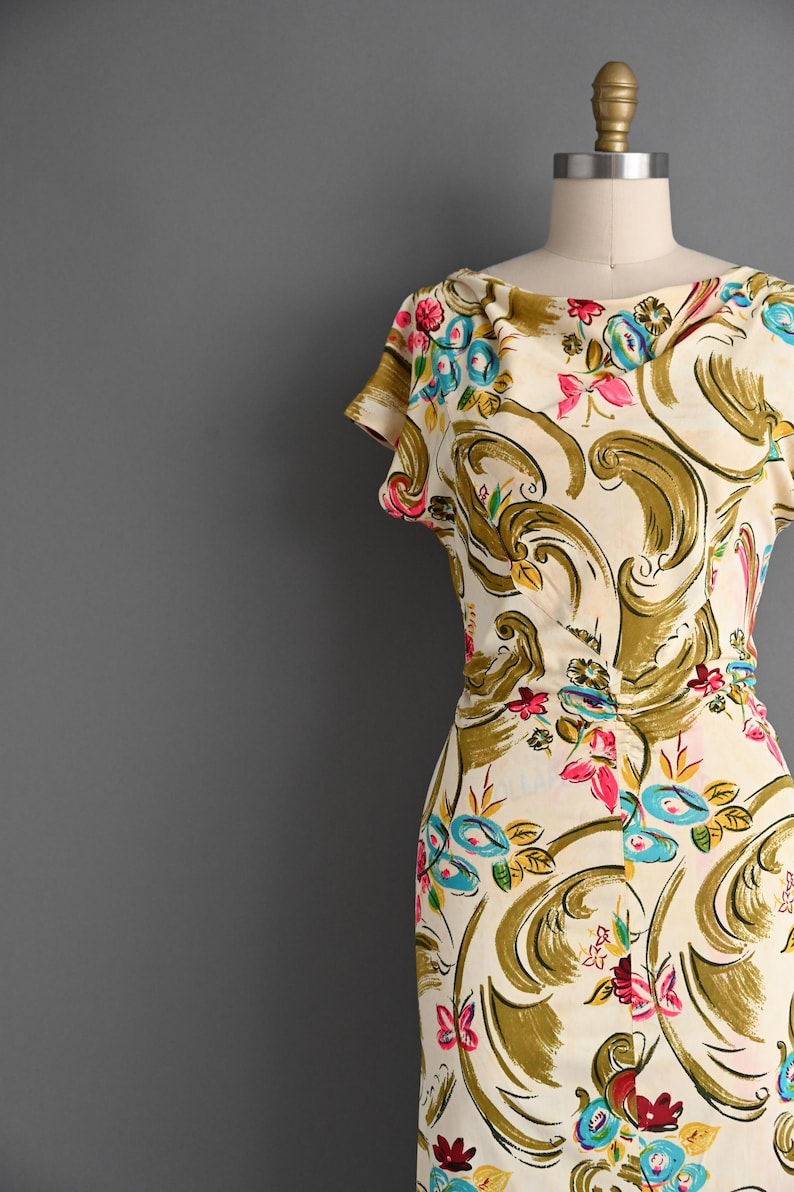 May include: A cream-colored dress featuring a cowl neckline and short sleeves, decorated with a repeating pattern of large olive green swirls, bright pink flowers, and small blue floral accents throughout the fabric.