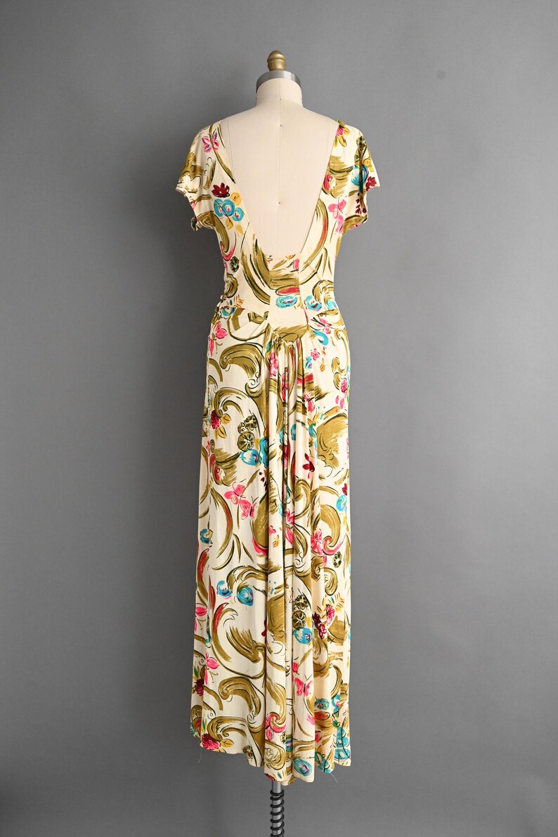 May include: A vintage-style, floor-length dress on a mannequin. The dress features a cream-colored background with a floral pattern in pink, blue, and gold. It has short sleeves and a low back.