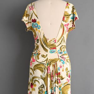 May include: A cream-colored dress featuring a vibrant floral and scroll pattern in shades of olive green, bright pink, and turquoise blue. The garment has short sleeves, a deep V-shaped back neckline, and gathered fabric at the waist.