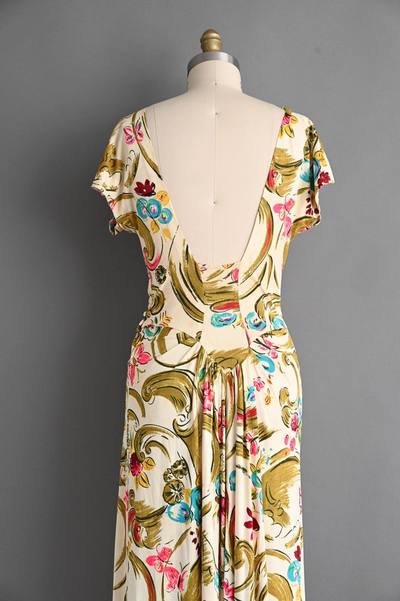 May include: A cream-colored dress featuring a vibrant floral and scroll pattern in shades of olive green, bright pink, and turquoise blue. The garment has short sleeves, a deep V-shaped back neckline, and gathered fabric at the waist.