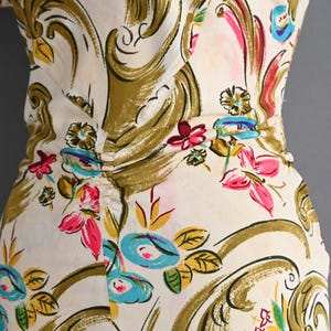 May include: A cream-colored dress featuring a large-scale olive green scroll pattern interspersed with vibrant floral motifs in shades of hot pink, bright blue, and yellow. The garment includes a gathered waist detail at the center.