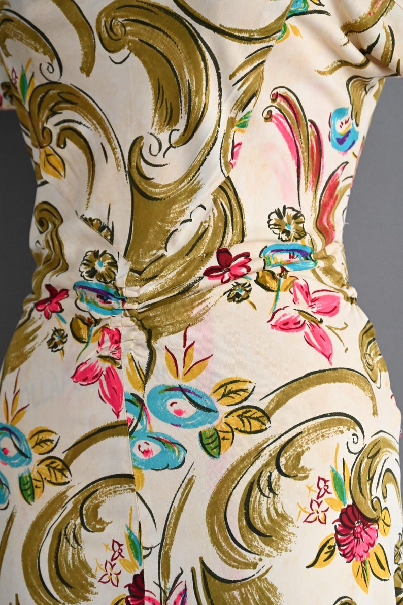 May include: A cream-colored dress featuring a large-scale olive green scroll pattern interspersed with vibrant floral motifs in shades of hot pink, bright blue, and yellow. The garment includes a gathered waist detail at the center.