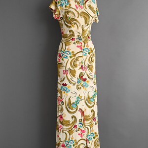 May include: A vintage ivory-colored dress with a floral pattern in shades of pink, blue, and green. The dress features short sleeves, a fitted waist, and a floor-length skirt with a front slit. The dress is displayed on a mannequin.