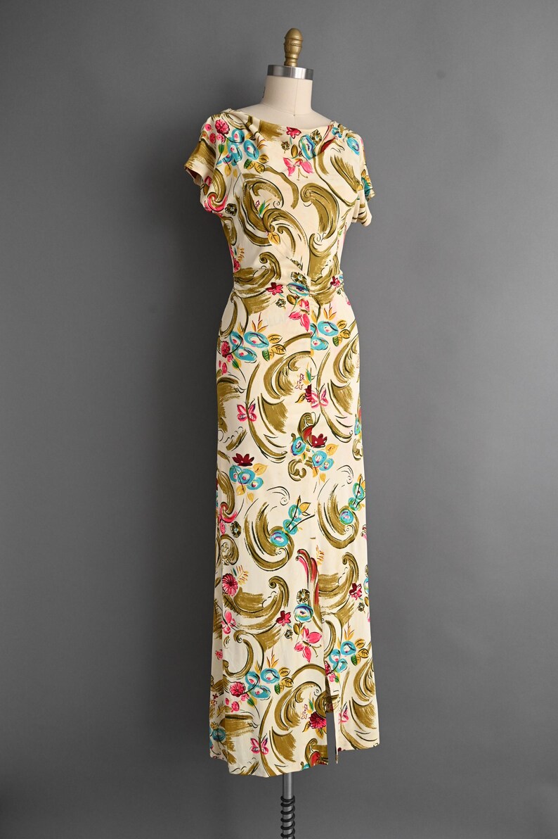 May include: A vintage ivory-colored dress with a floral pattern in shades of pink, blue, and green. The dress features short sleeves, a fitted waist, and a floor-length skirt with a front slit. The dress is displayed on a mannequin.