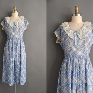 May include: A vintage blue and white plaid dress with a white lace collar and cap sleeves. The dress is displayed on a mannequin and has a gathered waist and tiered skirt. The dress is a midi length.