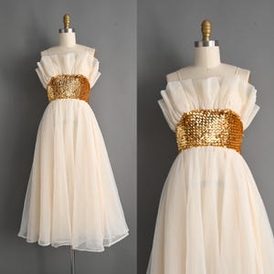 May include: A cream-colored dress with a gold sequined belt. The dress features a ruffled neckline and a flowing skirt. The dress is displayed on a mannequin against a gray background. The dress appears to be vintage or retro style.