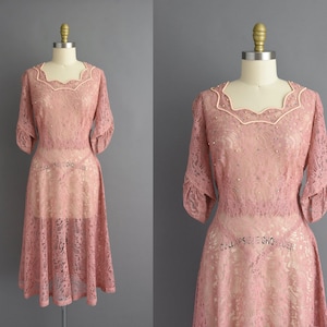 May include: A blush pink lace dress with a scalloped neckline and short sleeves. The dress has a fitted bodice and a full skirt.
