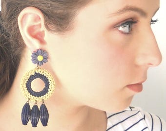 May include: A pair of dangle earrings with a crocheted circle design in yellow and blue with a small purple flower charm. The earrings have a fringe of dark blue beads.