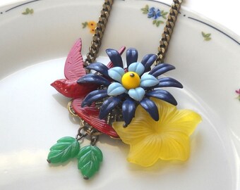 Puede incluir: Un collar colorido con una flor azul, amarilla y blanca, dos pájaros rojos y hojas verdes. El collar está hecho de metal y tiene una cadena dorada.
