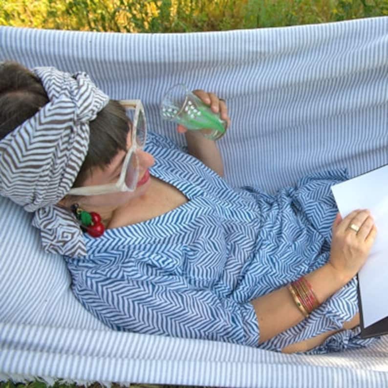 May include: A person wearing a blue and white patterned shirt and a gray and white patterned headband is relaxing in a hammock. They are holding a glass of green liquid and reading a book.