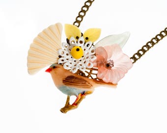 Puede incluir: Un collar de cadena dorada con un diseño de pájaro, flor y hoja. El pájaro es marrón claro con detalles azules y un pico rojo. Las flores son rosas y blancas. Las hojas son verdes.