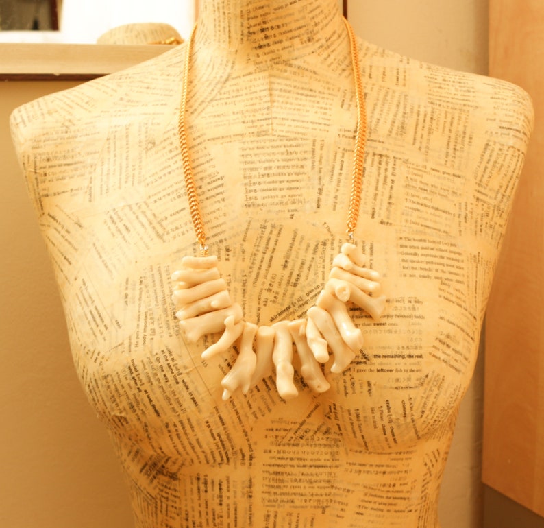 May include: A white coral necklace with a gold chain. The necklace is displayed on a mannequin with a patterned background.