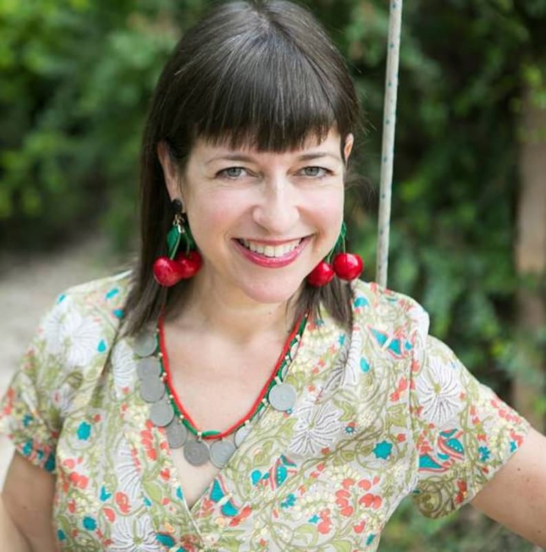 May include: A woman with dark brown hair wearing a floral print dress with a red and green beaded necklace and cherry earrings.