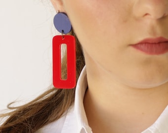 May include: Red and blue geometric earrings. The earrings are made of acrylic and have a rectangular shape with a smaller rectangle cut out in the center. The earrings are hanging from a silver post.