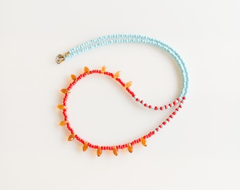 May include: A handmade necklace featuring a combination of red, orange, and light blue beads. The necklace has a gold-colored clasp and a unique design with orange teardrop-shaped beads along the red portion.