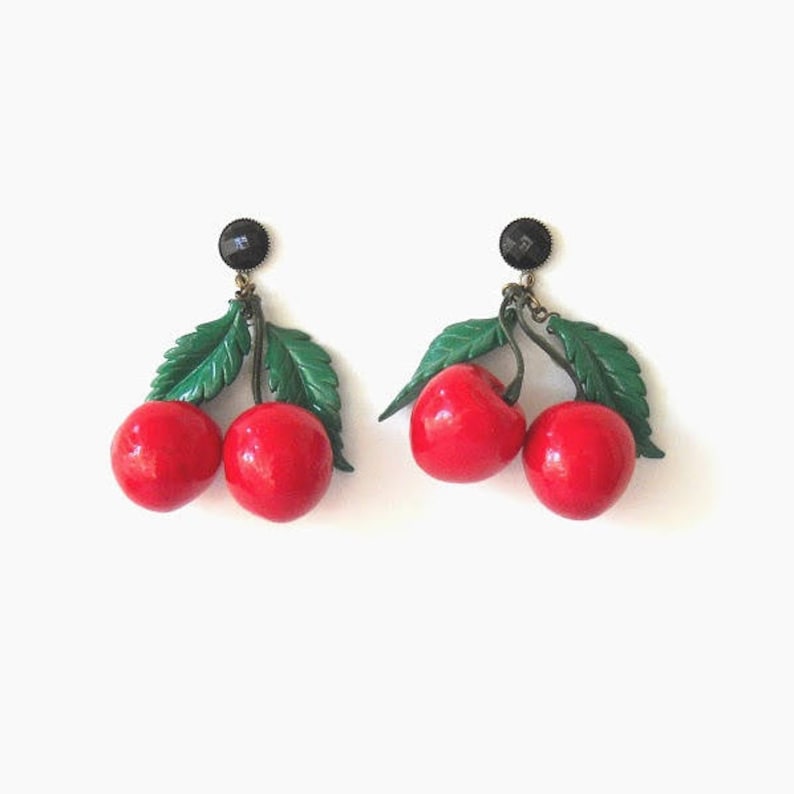 May include: A pair of dangle earrings featuring two red cherries with green leaves on each earring. The cherries are attached to a black post with a gold chain.