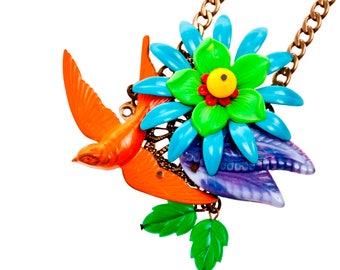 Puede incluir: Un collar colorido con un pájaro naranja brillante en vuelo, una flor grande con pétalos azules y verdes y hojas verdes. El collar tiene una cadena dorada. La flor tiene un centro amarillo con un acento rojo.