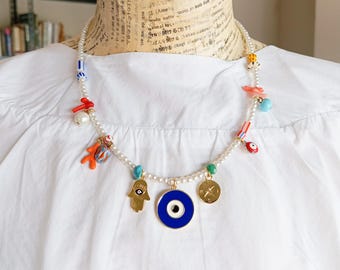 Könnte beinhalten: Eine Perlenkette mit bunten Anhängern. Sie enthält eine goldene Hamsa-Hand, ein blaues böses Auge und einen goldenen Münzanhänger. Weitere Anhänger sind Korallen, Perlen und ein kleiner roter Augenanhänger.