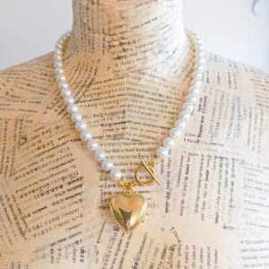 Può includere: Una collana di perle bianche con un ciondolo a forma di cuore dorato e una chiusura a scorrimento dorata. La collana è esposta su un manichino.