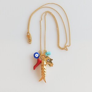 Puede incluir: Collar de cadena dorada con un dije de pez dorado, un chile rojo, un dije de ojo turco azul y una mano turquesa con un dije de ojo turco.