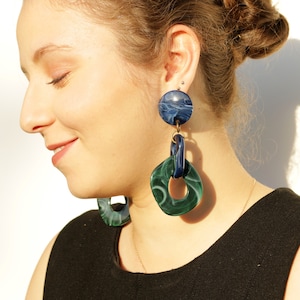 May include: A pair of statement earrings with a blue and green marbled design. The earrings feature a large round top piece and a smaller square piece with a ring in the center.