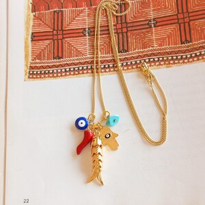 Puede incluir: Un collar de cadena dorada con un dije que presenta un pez, un chile rojo, un ojo azul malvado y una mano de hamsa turquesa.