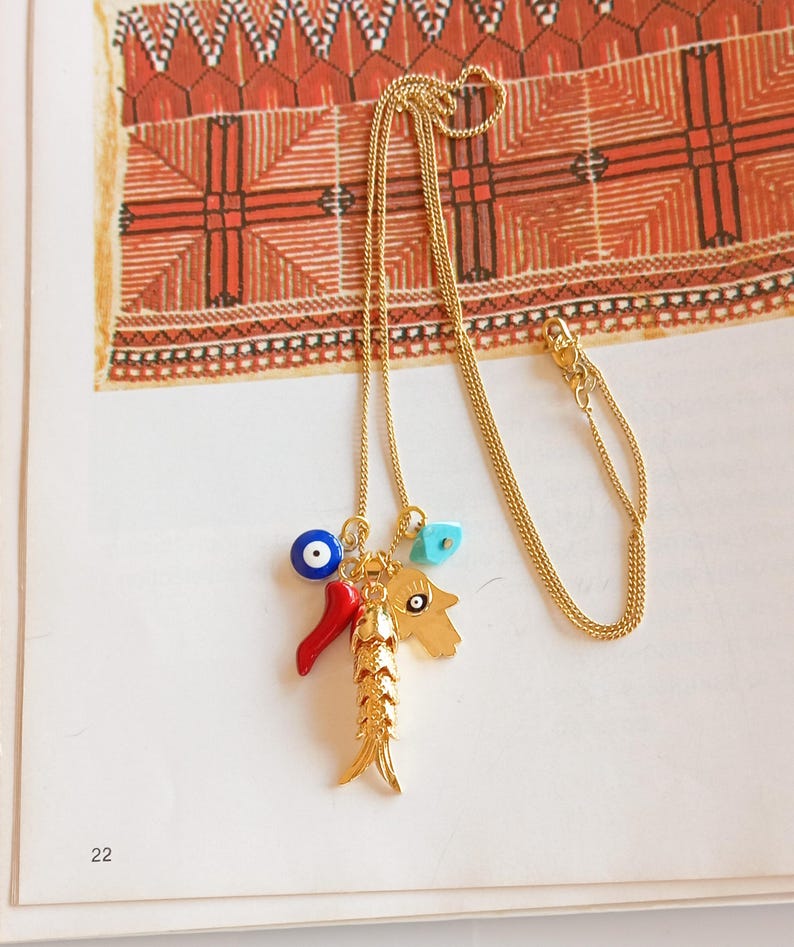 Puede incluir: Un collar de cadena dorada con un dije que presenta un pez, un chile rojo, un ojo azul malvado y una mano de hamsa turquesa.