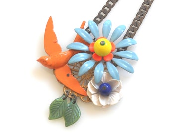 Puede incluir: Un collar colorido con un colgante de pájaro naranja, margarita azul y flor blanca. El colgante tiene abalorios de hojas verdes y una cadena marrón. Una pieza de joyería única y artesanal.