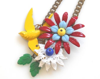 Puede incluir: Un collar con un dije de pájaro amarillo, flores rojas, azules y blancas y hojas verdes.