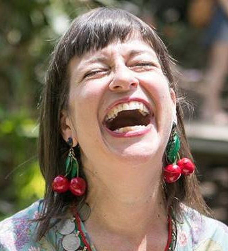 May include: A woman with brown hair and a bright smile is wearing a floral shirt and cherry earrings. The earrings are red and green and have a realistic cherry design.