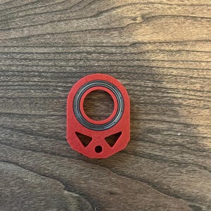 May include: A red, geometric-shaped object with a central circular opening. The object has a black and silver bearing visible within the opening. The design includes a small hole and two triangular cutouts. The item is likely a component or part.