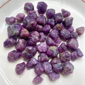 May include: A collection of small, raw, unpolished gemstones in varying shades of deep purple and magenta. These irregular, textured stones have a matte finish and are clustered together, displaying natural facets and earthy inclusions.