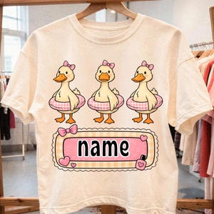 May include: Cream-colored t-shirt featuring three cartoon ducks wearing pink bows and inflatable rings. Each duck has orange feet and a yellow beak. Below the ducks is a pink and yellow banner with the word "name" in black.