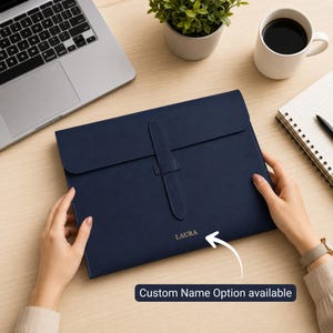 May include: A navy blue document folder with a flap closure and a vertical strap detail. The front of the folder features the name LAURA embossed in gold lettering. The folder is rectangular with a smooth finish and a secure central strap.