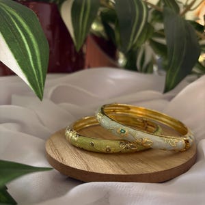 Peut inclure: Deux bracelets dorés avec des motifs floraux en émail vert clair et blanc, posés sur un petit disque en bois. Les bracelets ont des accents dorés et une petite pierre turquoise. Le fond comprend de la verdure et un tissu blanc.