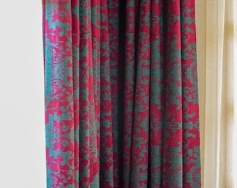 May include: Red and teal patterned curtains. The curtains feature a repeating floral design in teal against a red background. The fabric is draped in vertical folds, creating a textured appearance. The curtains are hanging in front of a white wall and a cream-colored sheer curtain.