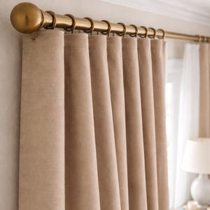 Pode incluir: Close-up de um conjunto de painéis de cortina bege pendurados numa barra dourada com anéis a condizer e um remate decorativo. As cortinas são feitas de um tecido espesso e texturizado e estão parcialmente corridas.