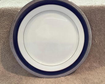 Op de afbeelding: Een witte keramische plaat met een marineblauwe band en een zilveren rand. De plaat heeft een dunne zilveren lijn in de buurt van het midden. Dit servies is geschikt voor dagelijks gebruik of speciale gelegenheden.