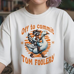 May include: White t-shirt with the text "Off to commit Tom Foolery". The graphic shows a raccoon in a cowboy hat riding a goose. The raccoon wears a vest and boots. The text and graphic are in orange.