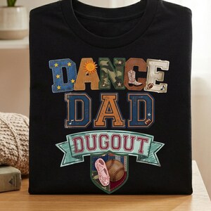 Könnte beinhalten: Schwarzes T-Shirt mit dem Schriftzug "DANCE DAD DUGOUT" in bunten Buchstaben im Patchwork-Stil. Das Design beinhaltet eine Sonne, einen Cowboy-Stiefel, einen Football, einen Ballettschuh und einen Baseball.