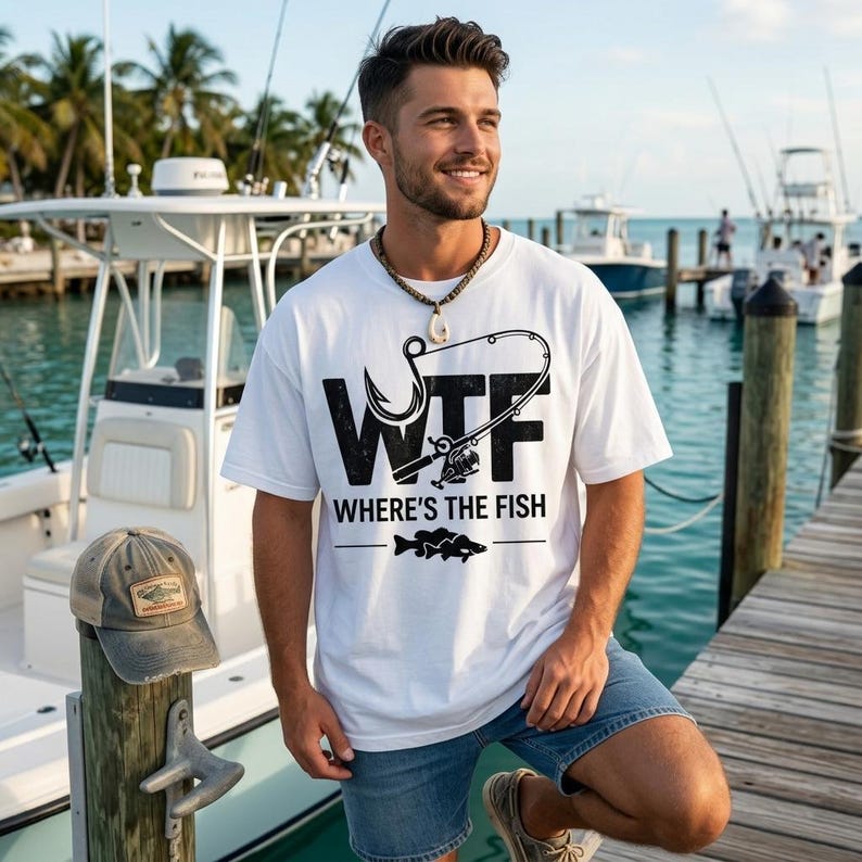 May include: White t-shirt with a black graphic that says "WTF WHERE'S THE FISH" and a fishing hook and rod design. A weathered gray and brown baseball cap rests on a wooden post. The background shows boats and water.