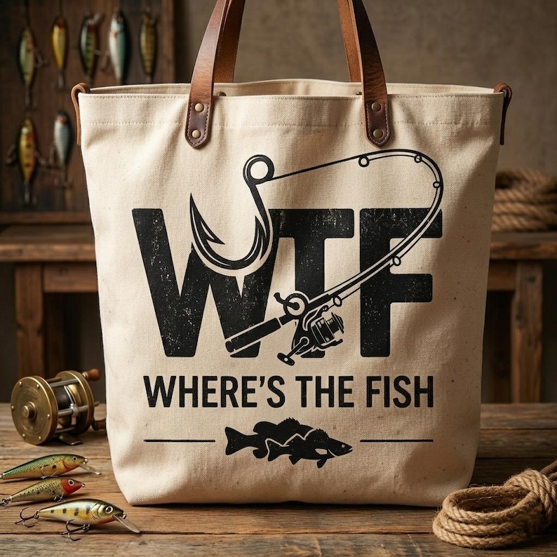 May include: A beige canvas tote bag with brown leather handles. The bag features a black graphic of a fishing hook and rod, with the letters "WTF" and the phrase "WHERE'S THE FISH" printed on it. A fish silhouette is below the text.