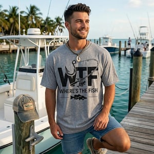 May include: A gray t-shirt with the text "WTF Where's the Fish" and a fishing hook and rod graphic. The image also shows a weathered baseball cap with a fishing-themed patch. The scene is set on a wooden dock with boats in the background.